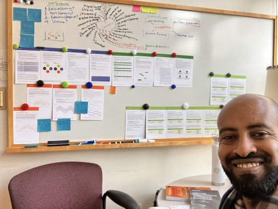 A person with dark complexion taking a selfie in front of a bulletin board.